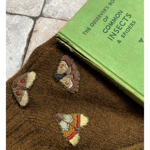 May include: A brown knitted jumper with three embroidered butterflies. The butterflies are in shades of brown, orange, yellow, and white. The jumper is lying on top of a green book titled "The Observer's Book of Common Insects & Spiders".
