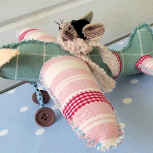 Puede incluir: Un avión de peluche con un cuerpo de tela a rayas rosa y blanco y una cola de tela a cuadros verde y blanco. Un pequeño ratón de peluche con pelaje marrón y blanco está sentado en la cabina.