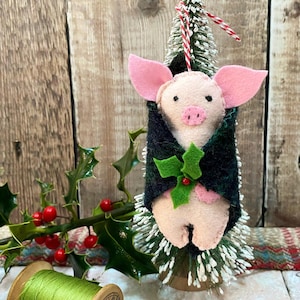 May include: A pink felt pig wearing a green and black plaid coat with a green holly sprig and red berry. The pig is hanging from a small white flocked Christmas tree.