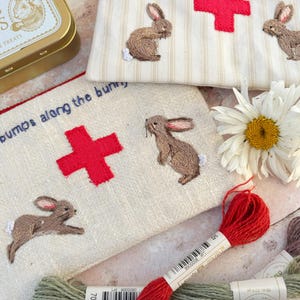 May include: Two beige fabric pouches featuring embroidered brown rabbits and red crosses. One pouch has the text "for when the whiskers wilt..." and the other "bumps along the bunny". Includes a gold tin, a white daisy, and embroidery floss.