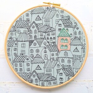 May include: Embroidered hoop art featuring a whimsical cityscape with multiple houses in black outline on a light blue background. The houses are arranged in a circular pattern and have various shapes and sizes. One house is highlighted in a peach color.