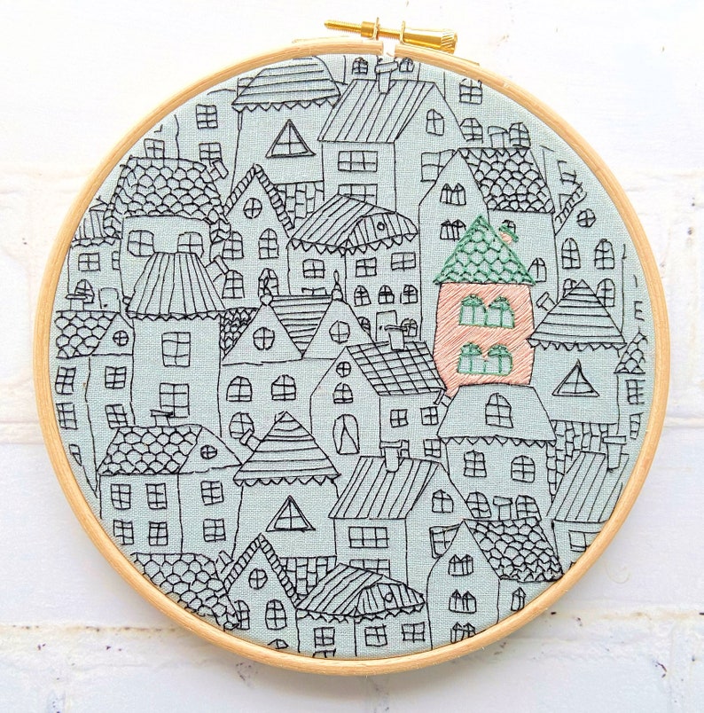 May include: Embroidered hoop art featuring a whimsical cityscape with multiple houses in black outline on a light blue background. The houses are arranged in a circular pattern and have various shapes and sizes. One house is highlighted in a peach color.