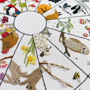 May include: Embroidered hoop art depicting a year-round calendar with animals and plants representing each month. The months are labeled around the edge of the hoop, with the months of August, October, November, and February visible in the image. The hoop features a variety of colors, including red, green, blue, yellow, and brown.