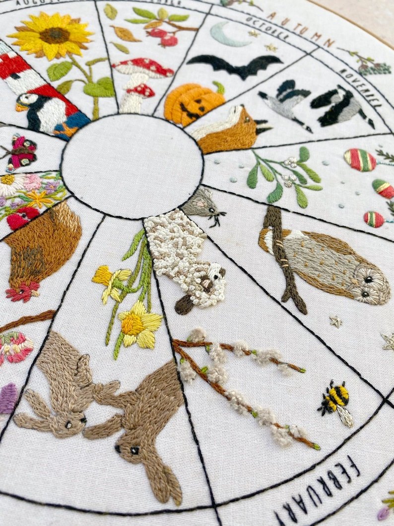 May include: Embroidered hoop art depicting a year-round calendar with animals and plants representing each month. The months are labeled around the edge of the hoop, with the months of August, October, November, and February visible in the image. The hoop features a variety of colors, including red, green, blue, yellow, and brown.