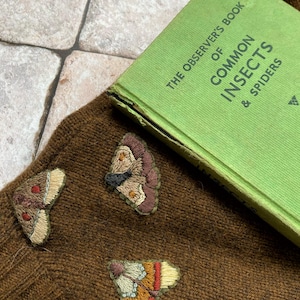 May include: A brown knitted sweater with three embroidered butterfly patches. The sweater is lying on a tiled surface with a green book titled "The Observer's Book of Common Insects & Spiders" on top of it.