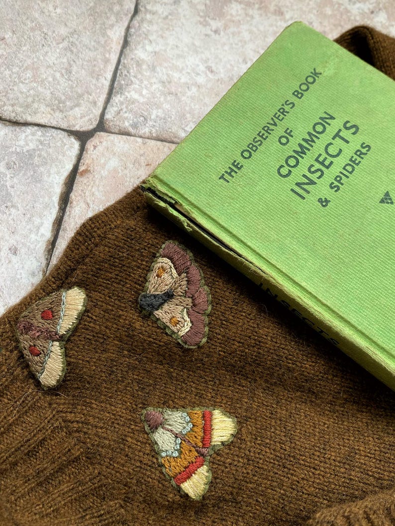 May include: A brown knitted sweater with three embroidered butterfly patches. The sweater is lying on a tiled surface with a green book titled "The Observer's Book of Common Insects & Spiders" on top of it.