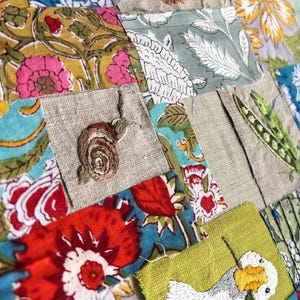 May include: A patchwork textile featuring squares of floral patterned fabric in red, yellow, blue, and green, interspersed with plain linen squares. Embroidered details include a brown snail, a green pea pod, and a white goose holding a flower.