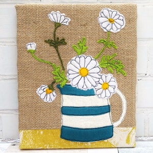 May include: A white and blue striped ceramic jug with a white daisy bouquet on a hessian background. The daisies are embroidered with white thread and have yellow centres.