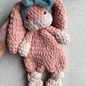 Könnte beinhalten: Ein gehäkeltes Plüsch-Hasenspielzeug aus weichem rosa Garn mit cremefarbener Schnauze, Füßen und Händen. Der Hase hat lange, hängende Ohren und trägt ein flauschiges, blaugrünes Stirnband auf der Stirn. Er hat kleine, dunkle, runde Augen.