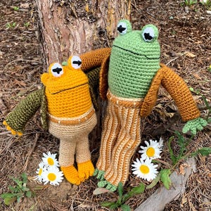 May include: Two crocheted frog plushies, one green and one orange, wearing brown and tan striped pants and green and brown jackets. The frogs are standing next to a tree trunk and some white daisies.