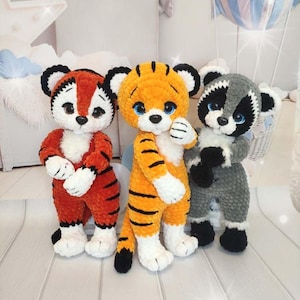 May include: Three handmade crochet plush toys standing side by side. The left toy is a red and white tiger with black stripes. The centre toy is an orange and white tiger with black stripes. The right toy is a grey and white raccoon with black markings.