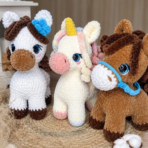 May include: Three plush toy horses are displayed. The white horse has brown accents and a blue flower. The unicorn is cream-colored with a pink nose and a yellow horn. The brown horse has a blue halter. All have large blue eyes.