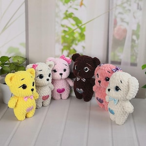 Może przedstawiać: Kolekcja ręcznie robionych szydełkowych misiów i psa w różnych kolorach. Misie są żółte, białe, różowe i brązowe, każdy z kokardką i sercem. Pies jest biały z niebieską kokardą. Ułożone są w rzędzie.