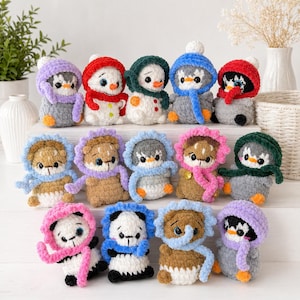 May include: A selection of handmade crochet plush toys, featuring penguins, snowmen, pandas, and bears. Each toy is adorned with a hat and scarf in shades of red, blue, pink, and purple. The toys are displayed on a white surface.