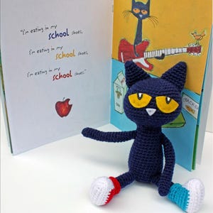 Puede incluir: Un juguete de peluche de gato azul tejido a crochet sentado en una superficie blanca con un libro abierto detrás. El libro tiene un gato negro con una guitarra roja en la portada. El texto del libro dice "Estoy comiendo en mis zapatos de la escuela, estoy comiendo en mis zapatos de la escuela, estoy comiendo en mis zapatos de la escuela."