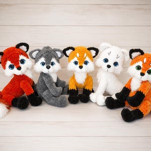 May include: Five plush stuffed animals made of soft yarn, including two orange and white foxes with black ears and feet, one gray wolf, one orange fox with black accents, and one white animal, all featuring large, round, glossy eyes.