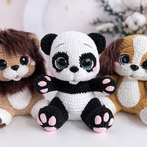 Op de afbeelding: Drie gehaakte knuffels: een leeuw, een panda en een hond. De leeuw is bruin met een bruine manen, de panda is zwart en wit, en de hond is bruin en wit. Elk heeft grote, expressieve ogen.