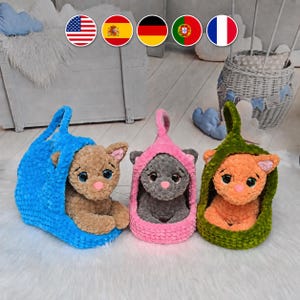 May include: Three plush cat toys, each nestled in a matching crocheted hooded basket. The left cat is tan in a blue basket, the centre is grey in a pink basket, and the right is orange in a green basket. All have large eyes and pink noses.