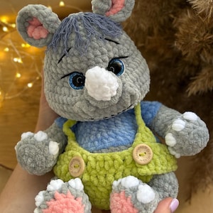 May include: A handmade gray crocheted rhino plush toy with blue eyes, pink inner ears, and a white snout. It wears a blue shirt and green overalls with wooden buttons. The toy is soft and cuddly.