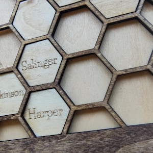 May include: Wooden honeycomb-shaped wall art with engraved names. The art piece features a natural wood finish with dark brown outlines. The names "Salinger" and "Harper" are visible. The design is reminiscent of a beehive.