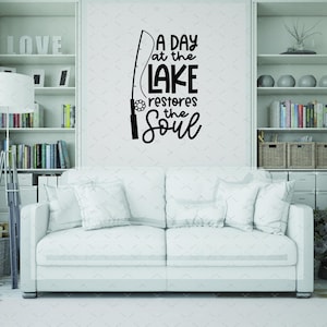 May include: A white sofa with several pillows is in front of a wall with a black decal. The decal reads "A day at the lake restores the soul" with a fishing rod graphic. Bookshelves are on either side.