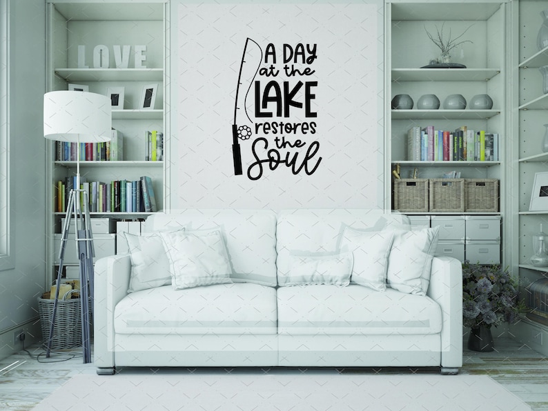 May include: A white sofa with several pillows is in front of a wall with a black decal. The decal reads "A day at the lake restores the soul" with a fishing rod graphic. Bookshelves are on either side.