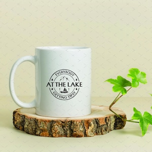 May include: White ceramic mug with a black circular design that reads "Everybody at the Lake Getting Tipsy." The mug sits on a wooden coaster with a green plant to the right. A perfect gift for lake lovers.