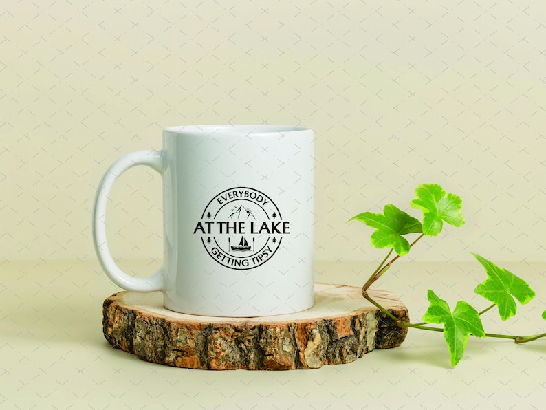 May include: White ceramic mug with a black circular design that reads "Everybody at the Lake Getting Tipsy." The mug sits on a wooden coaster with a green plant to the right. A perfect gift for lake lovers.