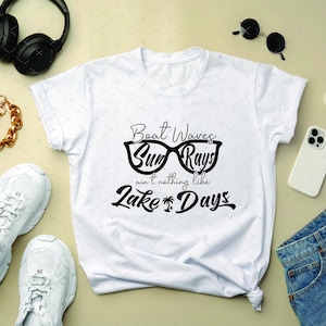 May include: White t-shirt with the text "Boat Waves, Sun Rays, ain't nothing like Lake Days" in black. The shirt is surrounded by accessories including white sneakers, a gold chain bracelet, black headphones, sunglasses, a phone, and denim shorts.