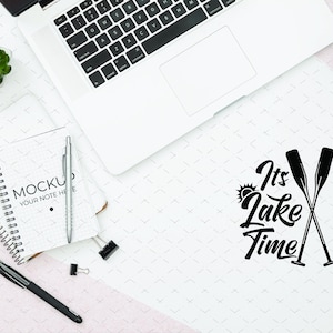 May include: Overhead shot of a workspace with a laptop, notebook, pens, and small plants. The notebook reads "Mockup Your Note Here." The phrase "It's Lake Time" is written in black script with crossed oars.