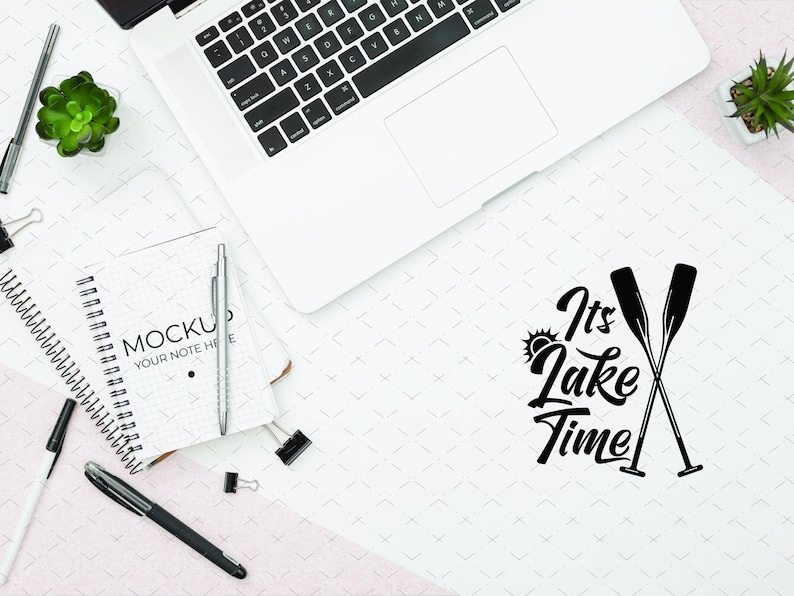 May include: Overhead shot of a workspace with a laptop, notebook, pens, and small plants. The notebook reads "Mockup Your Note Here." The phrase "It's Lake Time" is written in black script with crossed oars.
