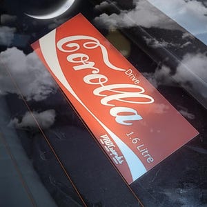 Puede incluir: Pegatina roja y blanca con el texto "Corolla Drive 1.6 Litre" y el logotipo "Pro-Works" en blanco. La pegatina está en una ventana de coche.