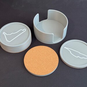 Puede incluir: Conjunto de seis posavasos grises con un contorno blanco de un circuito de carreras, y un portavasos gris. Un posavasos es de corcho.