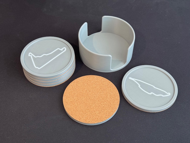 Puede incluir: Conjunto de seis posavasos grises con un contorno blanco de un circuito de carreras, y un portavasos gris. Un posavasos es de corcho.