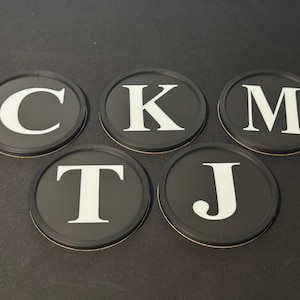 Puede incluir: Cinco posavasos redondos negros con letras mayúsculas blancas: C, K, M, T y J. Cada posavasos tiene un borde fino de color claro. Los posavasos están colocados sobre una superficie oscura.