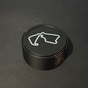 Puede incluir: Un juego de seis posavasos negros con un contorno blanco de un circuito de carreras en el posavasos superior. Los posavasos est&aacute;n apilados uno encima del otro.