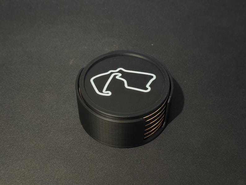 Puede incluir: Un juego de seis posavasos negros con un contorno blanco de un circuito de carreras en el posavasos superior. Los posavasos est&aacute;n apilados uno encima del otro.