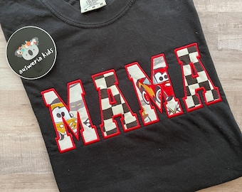 May include: Black t-shirt with a red and white checkered "MAMA" graphic featuring cartoon race cars. The graphic has red stitching around the letters.
