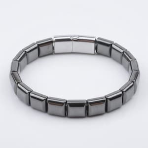 Peut inclure: Un bracelet gris foncé avec des perles carrées. Le bracelet est doté d'un fermoir argenté. Les perles sont disposées en cercle, créant un design élégant et moderne. Le bracelet est fait d'un matériau métallique brillant.