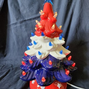 May include: A ceramic Christmas tree in red, white, and blue, with a blue star topper. The tree is lit with small, colorful lights. The base is red with white stars. A festive holiday decoration.