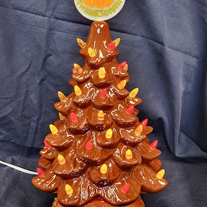 May include: A brown ceramic Thanksgiving tree with a pumpkin on top. The tree is decorated with lights and has a base with fall leaves and a turkey.