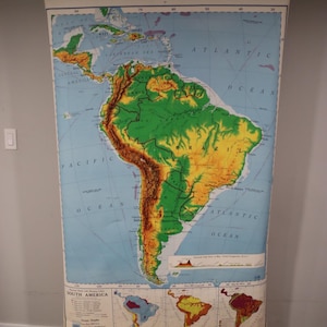 May include: A large, vertical wall-mounted pull-down map of South America. The map features topographic shading in green, yellow, and brown, with blue oceans. It includes smaller inset maps at the bottom, a legend, and the text South America.