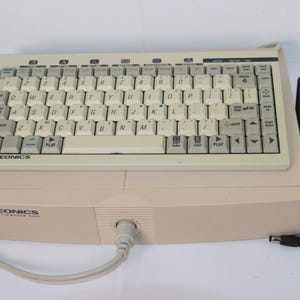 Puede incluir: Un dispositivo Videonics Video Titlemaker 3000 de color beige con teclado QWERTY integrado, teclas claras con letras negras, un cable en espiral y un adaptador de corriente negro independiente con cable. La marca está impresa en el frontal.