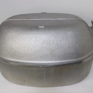 Puede incluir: Una fuente de horno ovalada grande de metal color plata con tapa a juego. El exterior de la tapa presenta un acabado texturizado y granulado, mientras que la base tiene una superficie lisa. Dos asas resistentes están fijadas a cada lado.