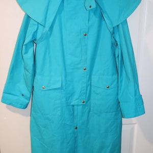 May include: A bright turquoise long coat featuring a wide shoulder cape, a high collar, and a front placket with gold-tone metal snap buttons. The coat includes two front flap pockets and a dark interior label with a small Australian flag graphic.