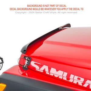 May include: A white vinyl decal featuring the word SAMURAI in a bold, stylized font applied to the red hood of a vehicle. The decal is positioned near the front corner, accompanied by a circular black and white premium quality seal.
