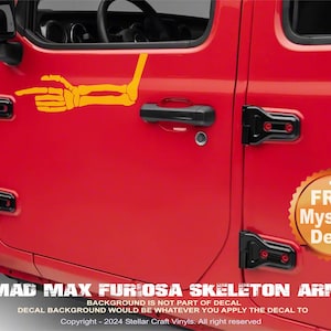 May include: A red Jeep Wrangler with a yellow skeleton arm decal on the driver's side door. The decal is pointing to the right. The text "MAD MAX FURIOSA SKELETON ARM" is printed below the decal. The text "+ FREE Mystery Decal" is printed on a yellow circle on the door.