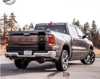May include: A dark gray pickup truck featuring a black vertical decal on the tailgate. The decal displays a circular Imperial crest emblem centered within a solid black stripe. The truck has a chrome rear bumper and multi-spoke alloy wheels.