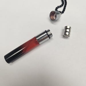 K&ouml;nnte beinhalten: Eine zerlegte Anh&auml;ngerhalskette mit einem zylindrischen Fl&auml;schchen. Das Fl&auml;schchen ist transparent mit einem Farbverlauf von Rot zu Schwarz. Die Halskette hat eine silberne Kappe und einen kleinen silbernen Magnetverschluss. Die Kette ist schwarz.