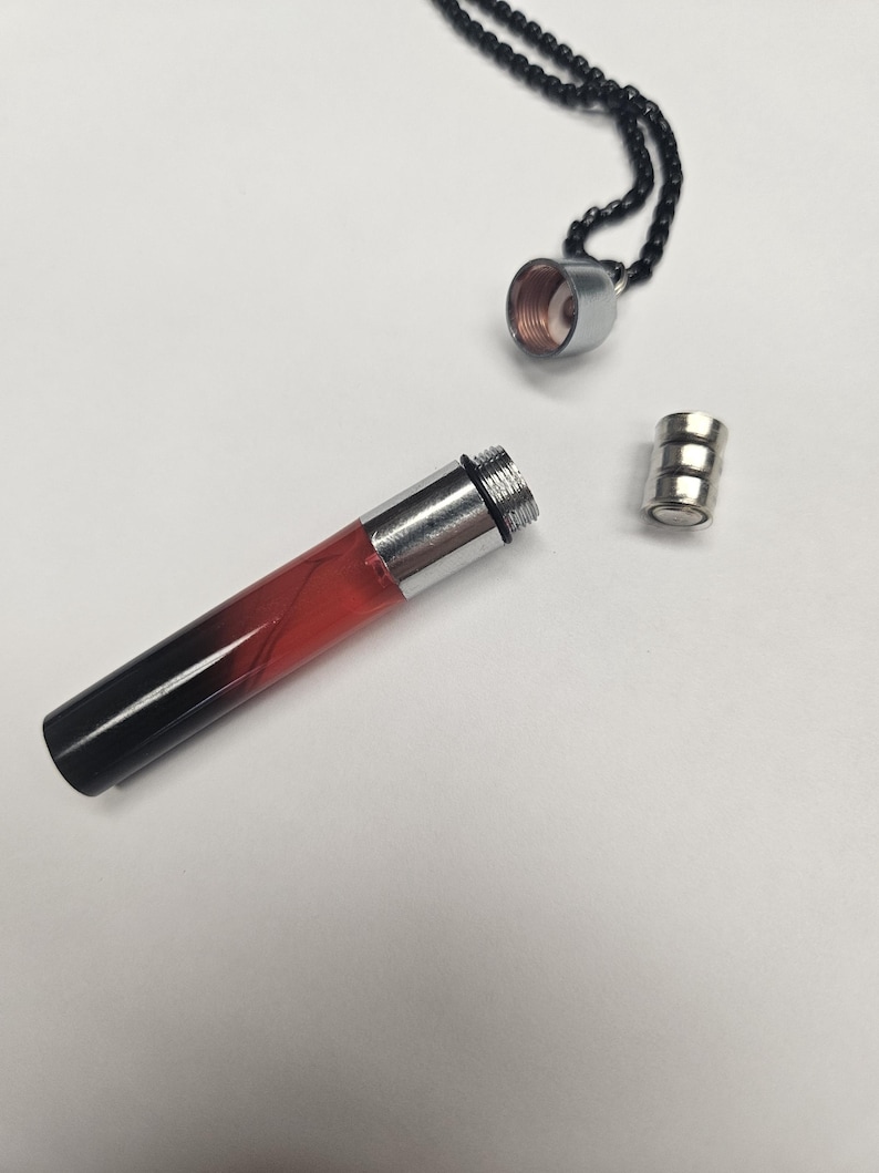 K&ouml;nnte beinhalten: Eine zerlegte Anh&auml;ngerhalskette mit einem zylindrischen Fl&auml;schchen. Das Fl&auml;schchen ist transparent mit einem Farbverlauf von Rot zu Schwarz. Die Halskette hat eine silberne Kappe und einen kleinen silbernen Magnetverschluss. Die Kette ist schwarz.